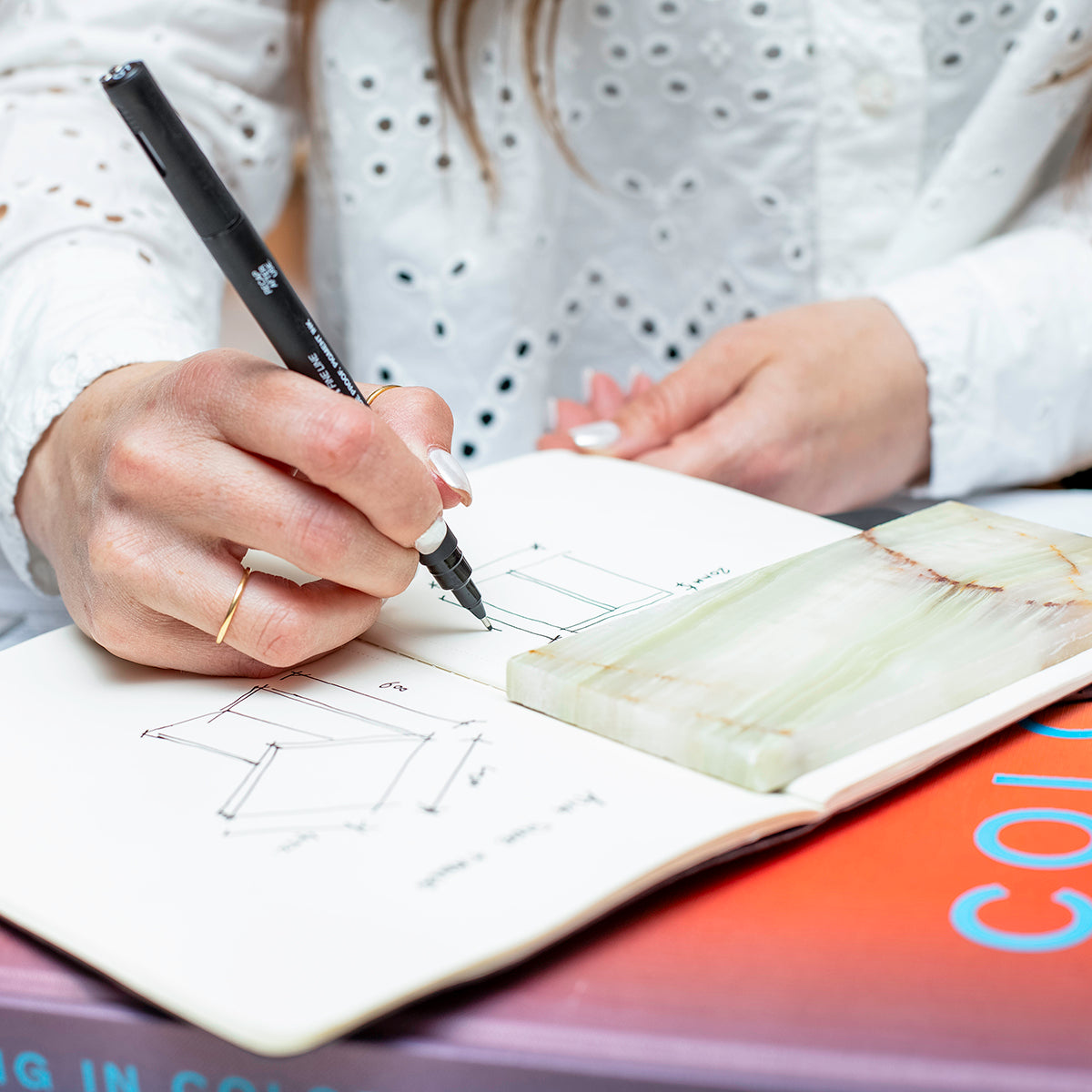 Close-up of a hand sketching a coffee table design in a sketch book with a marble swatch.