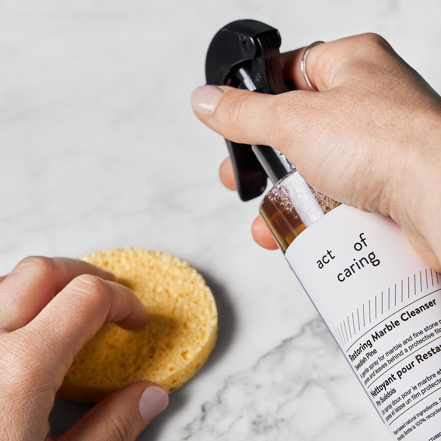 Hands holding  ‘Act of Caring’ Restoring Marble Cleanser and a round sponge.