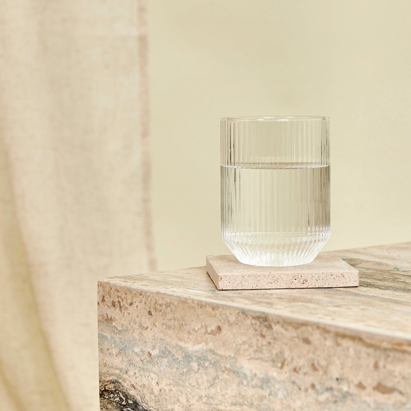 A detail of a glass of water and Travertine Marble coaster on marble side table.