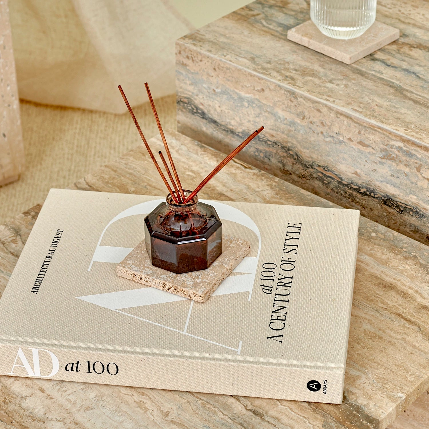 A close-up of a glass diffuser and travertine marble coaster and book on an Elba Silver travertine side table.