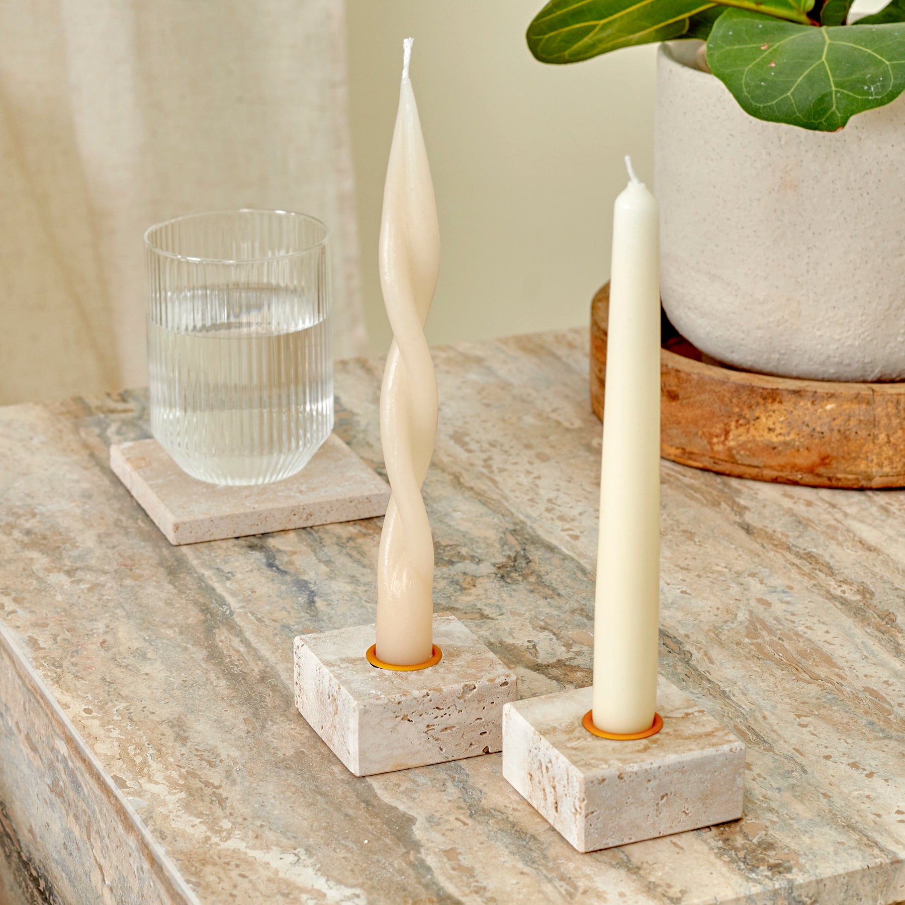 A close-up of two travertine marble candle holders with brass details and a glass of water on a travertine coaster, placed on a silver travertine, Elba side table.