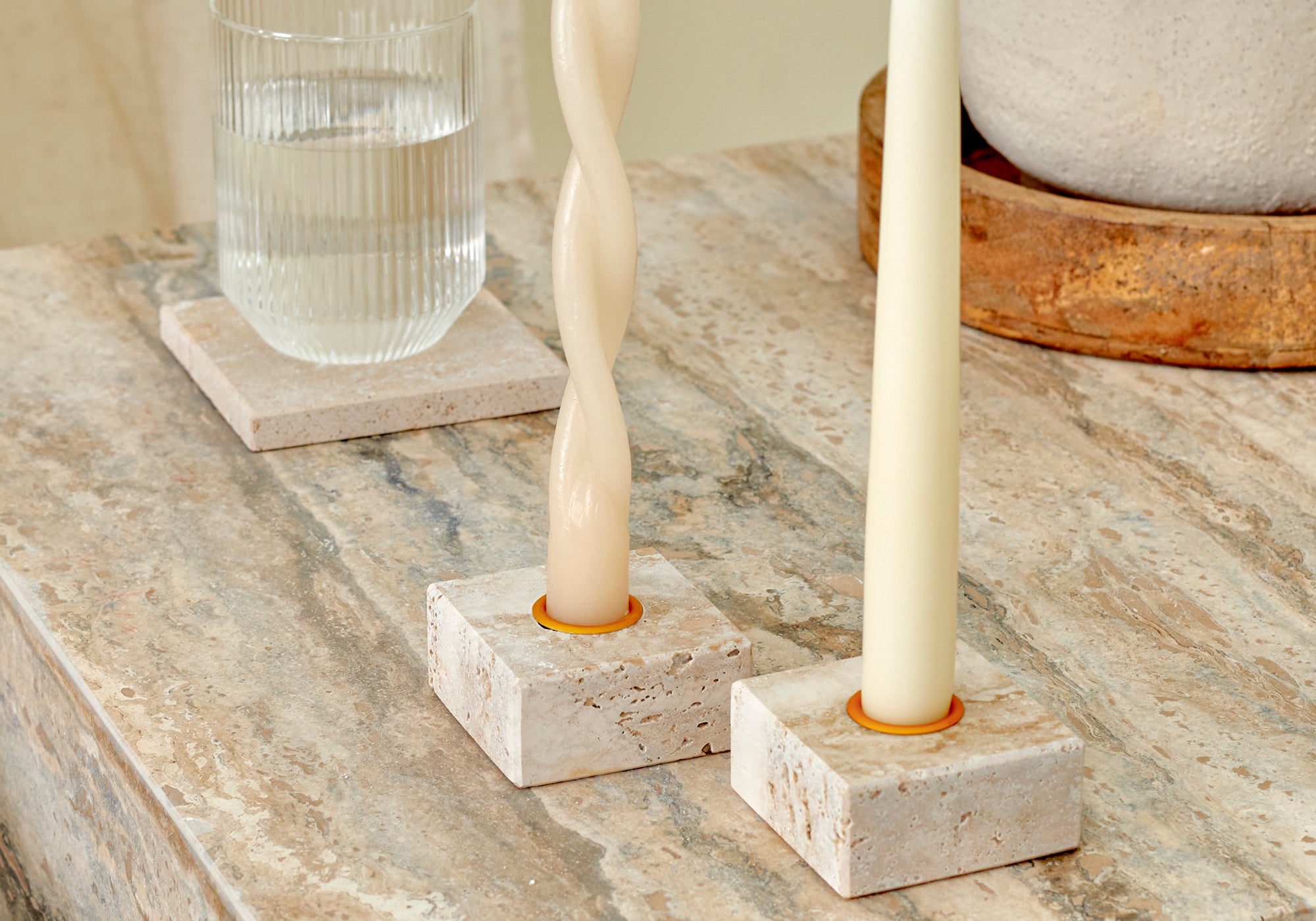 A close-up of two travertine marble candle holders with brass details and a glass of water on a travertine coaster, placed on a silver travertine, Elba side table.